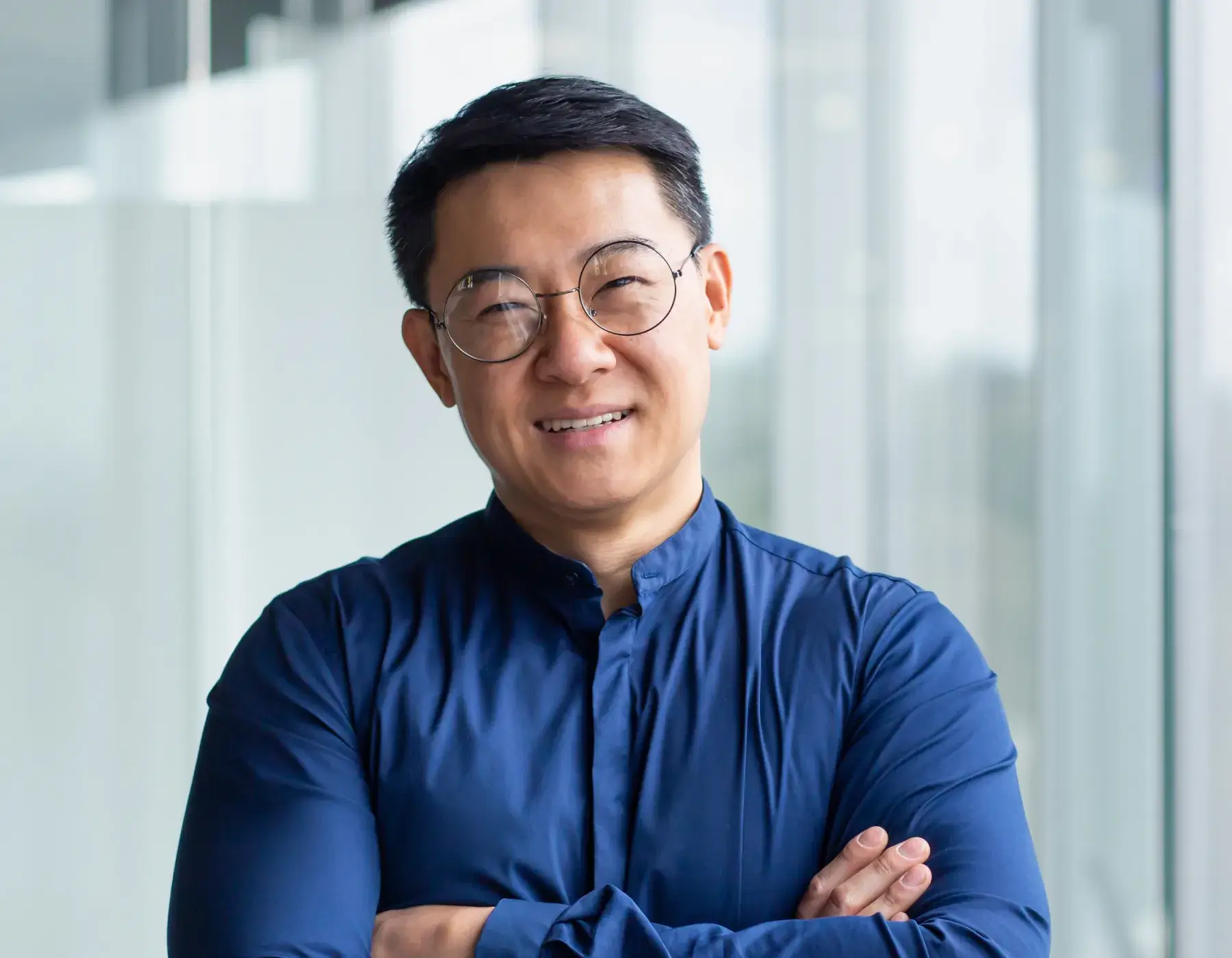 Smiling man with glasses and folded arms wearing a blue shirt in a bright modern office.