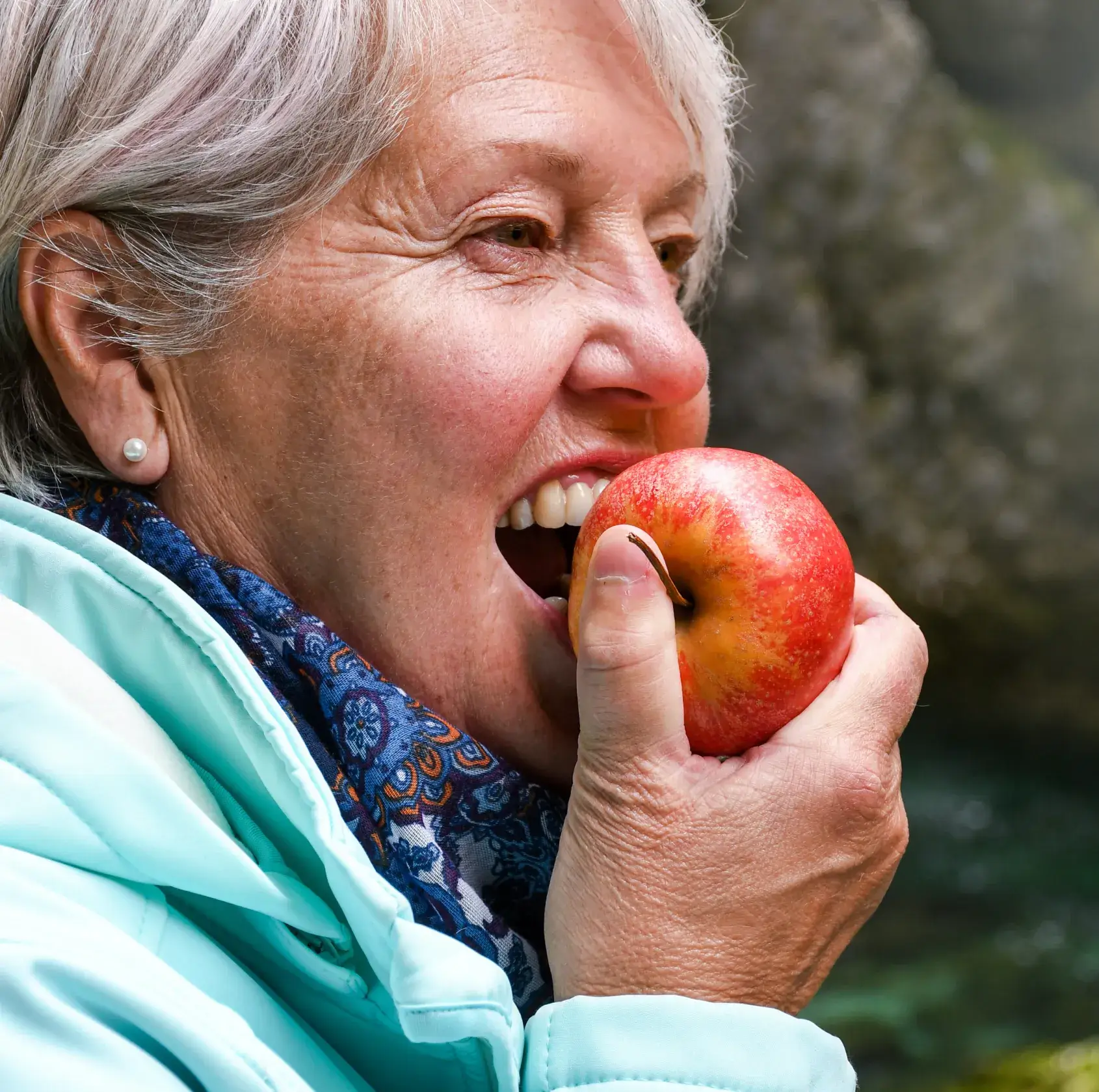 Older woman in a light jacket biting into a red apple outdoors.