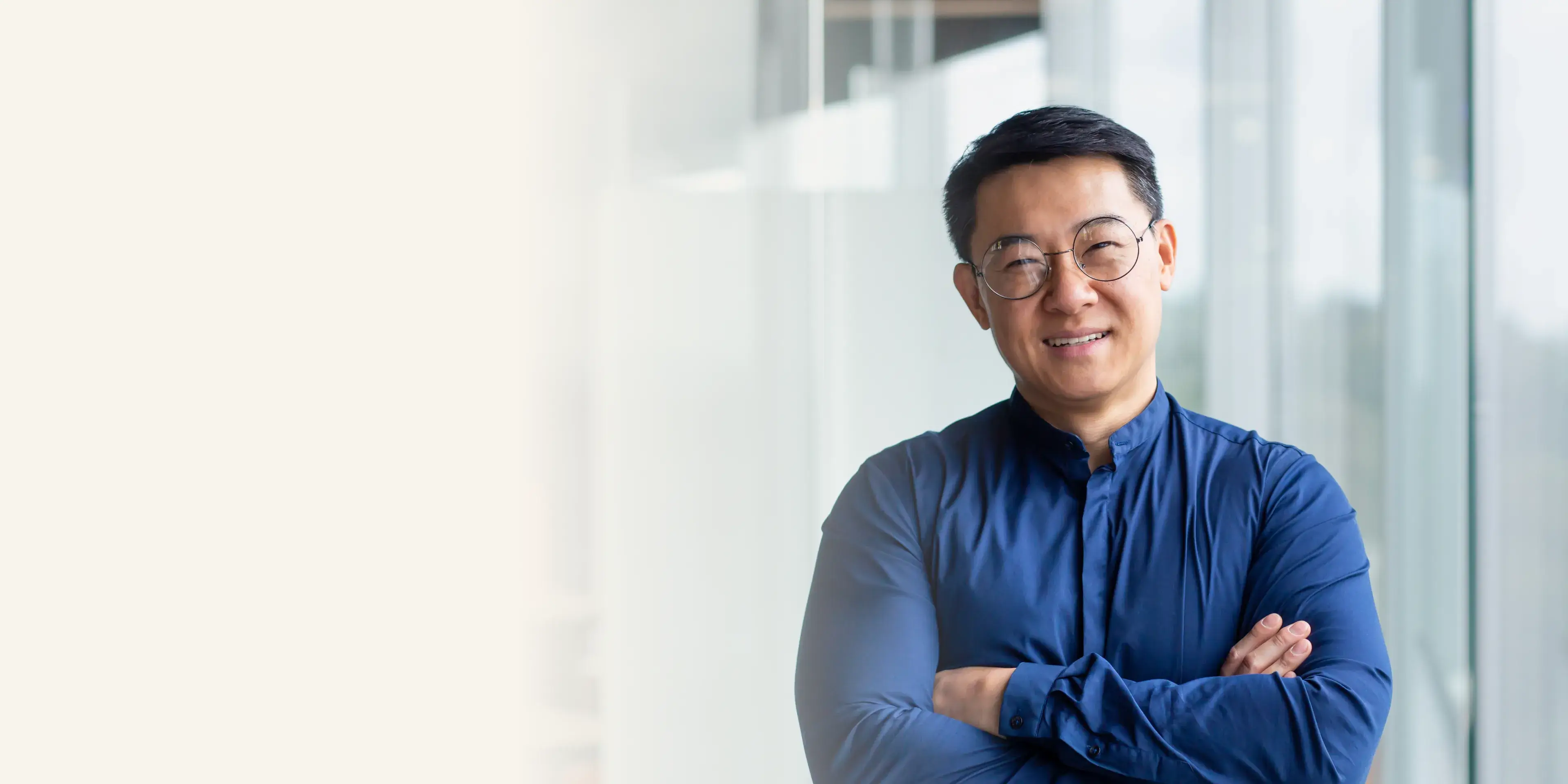 Smiling man with glasses and blue shirt standing with arms crossed by a window.