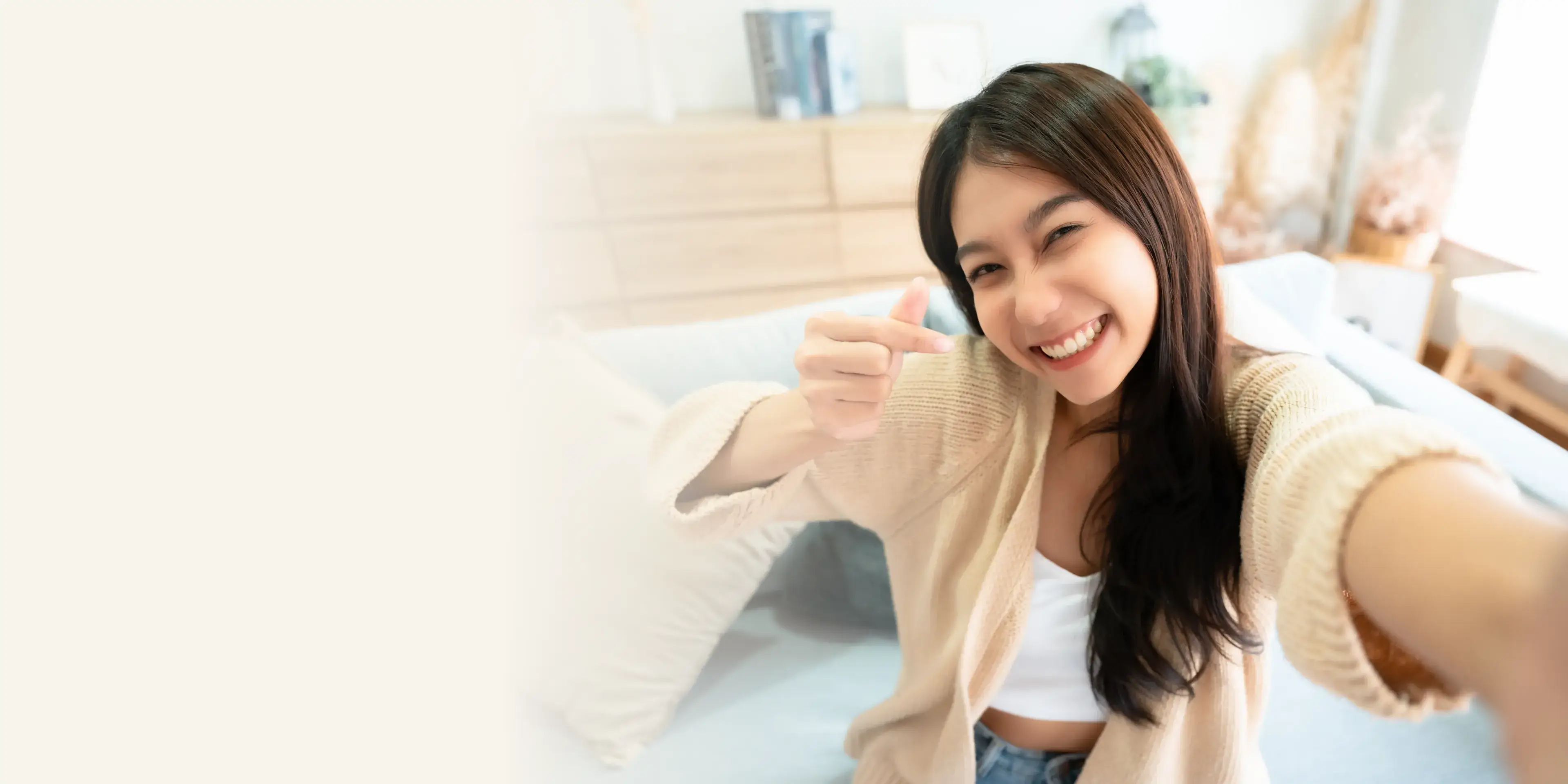 Smiling young woman making a heart gesture with fingers while taking a selfie indoors.